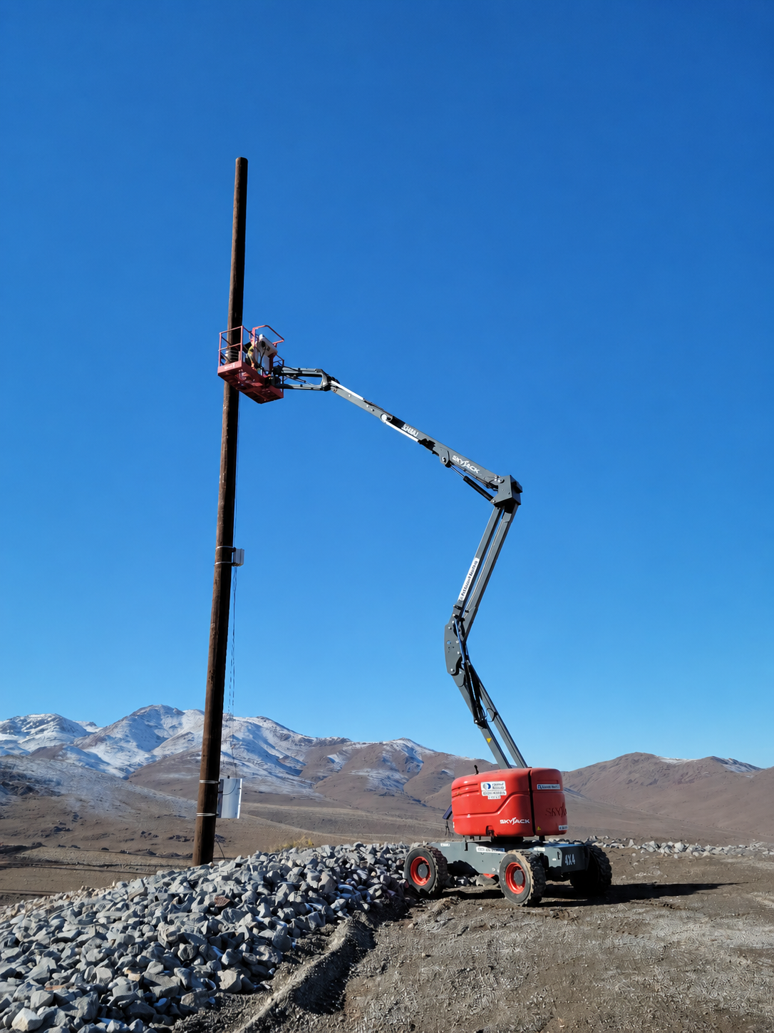 Technician on a boom lift installing equipment on a tall pole at a remote industrial site with snow-covered mountains nearby.