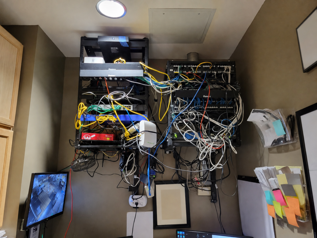 Chaotic wall-mounted technology racks with network cabling, power supplies, PoE injectors, and surveillance equipment at a newly onboarded customer site.