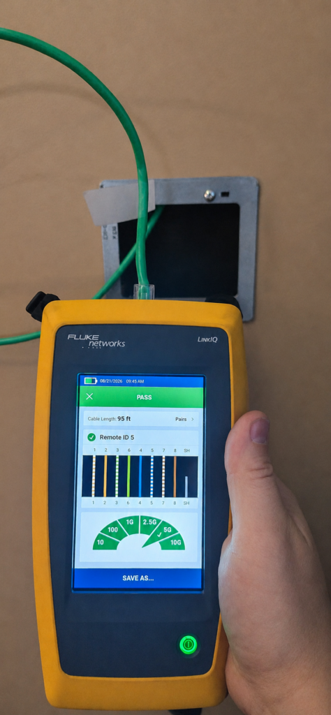 Handheld network cable tester showing a passing result on a green cable emerging from a wall box.