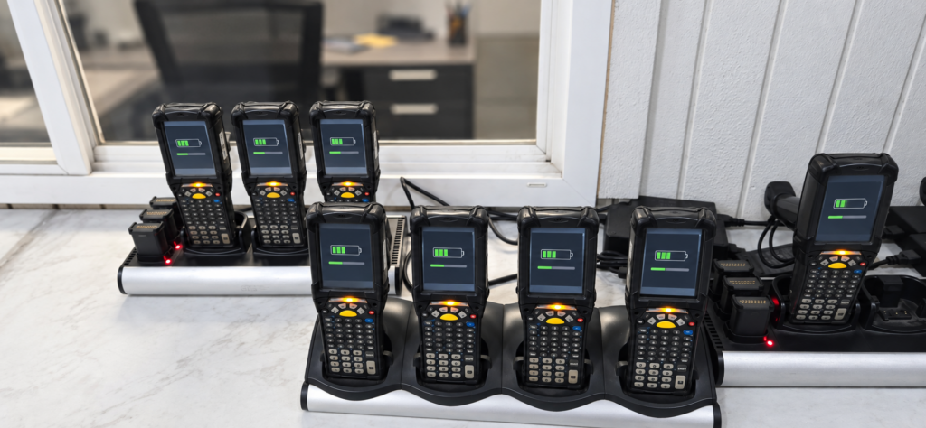 Rugged RF handheld scanners and spare batteries charging in multi-bay docks at a warehouse.