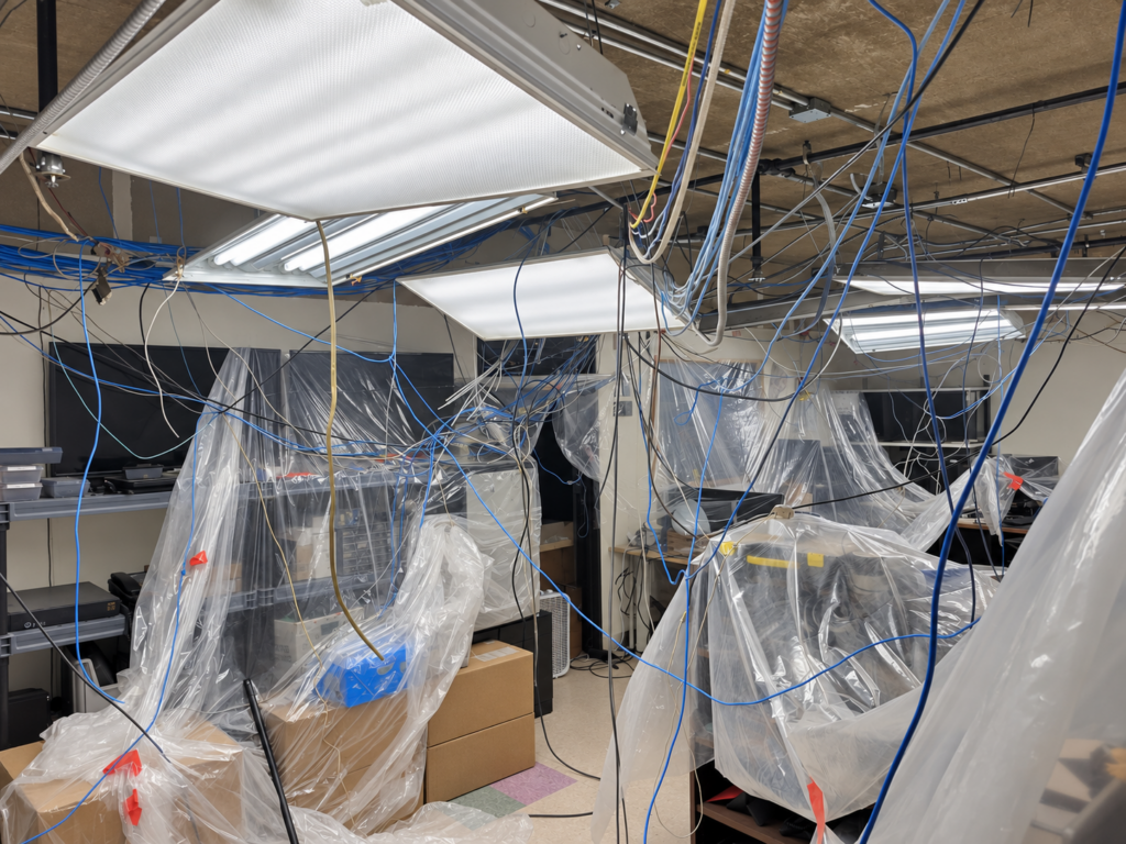 IT office with ceiling tiles removed, exposing years of unsupported low-voltage cabling above covered equipment and work areas.