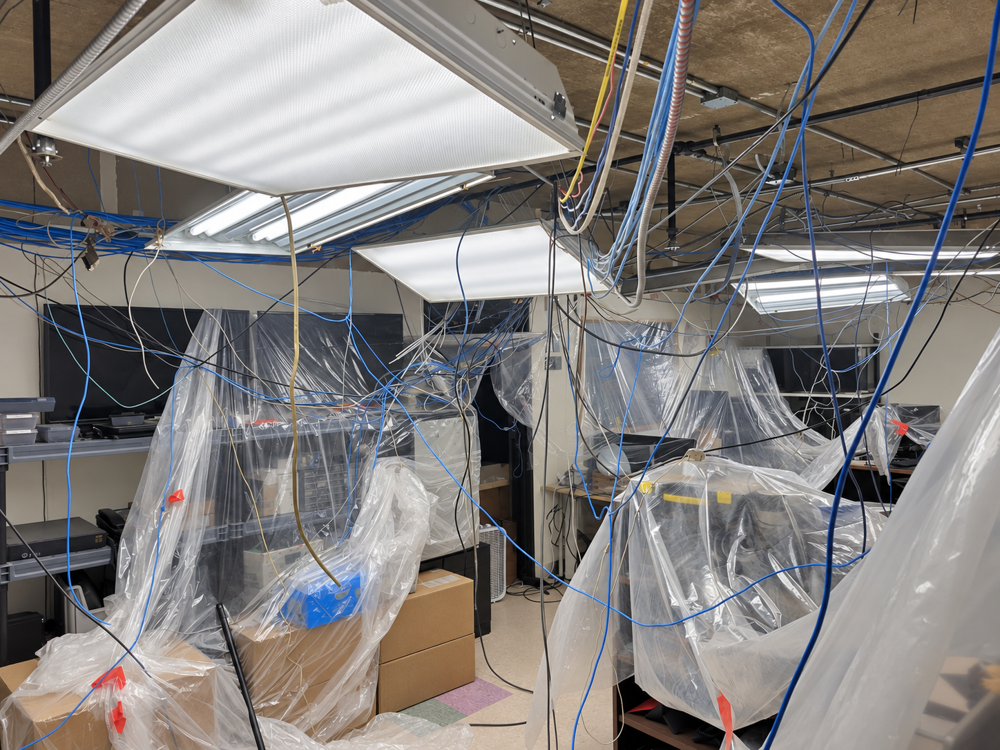 IT office with ceiling tiles removed, exposing years of unsupported low-voltage cabling above covered equipment and work areas.